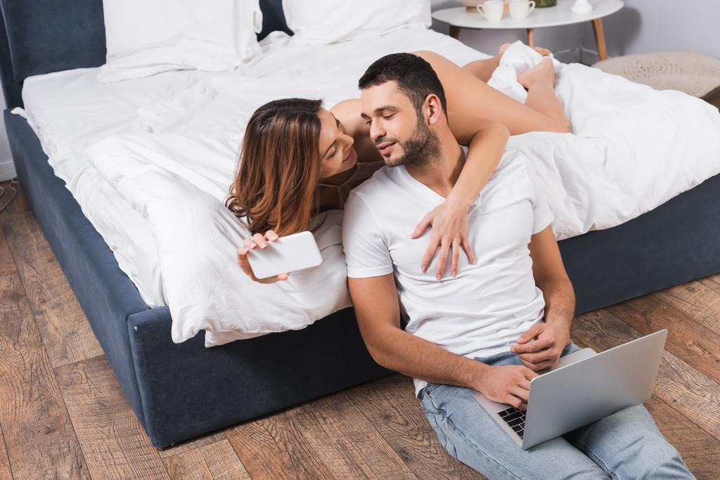 Smiling woman with smartphone hugging boyfriend using laptop in bedroom  - Photo, Image