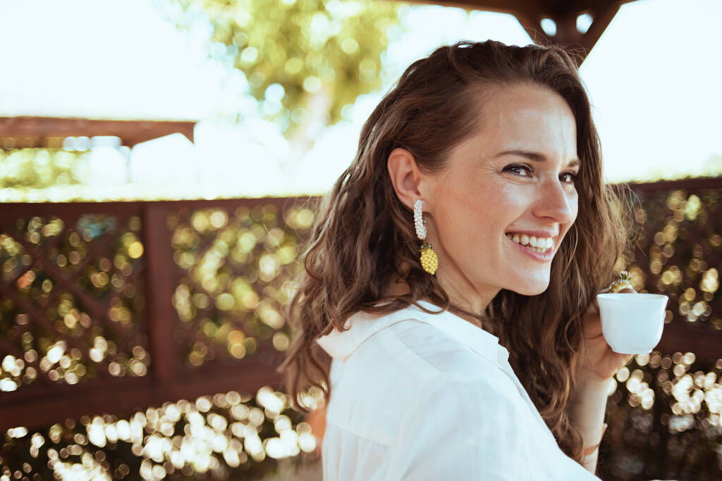 Portret uśmiechniętej młodej kobiety w białej koszuli z filiżanką kawy na tarasie. - Zdjęcie, obraz