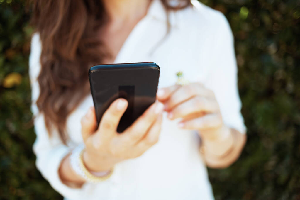 Szoros kép a fehér inges nő okostelefon alkalmazások a szabadban, közel a zöld fal. - Fotó, kép