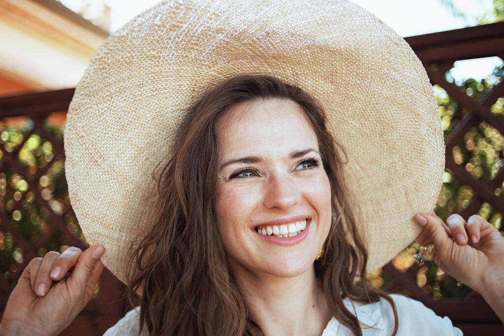 χαρούμενη μοντέρνα γυναίκα σε λευκό πουκάμισο με καπέλο στο αίθριο του ξενώνα ξενοδοχείο. - Φωτογραφία, εικόνα