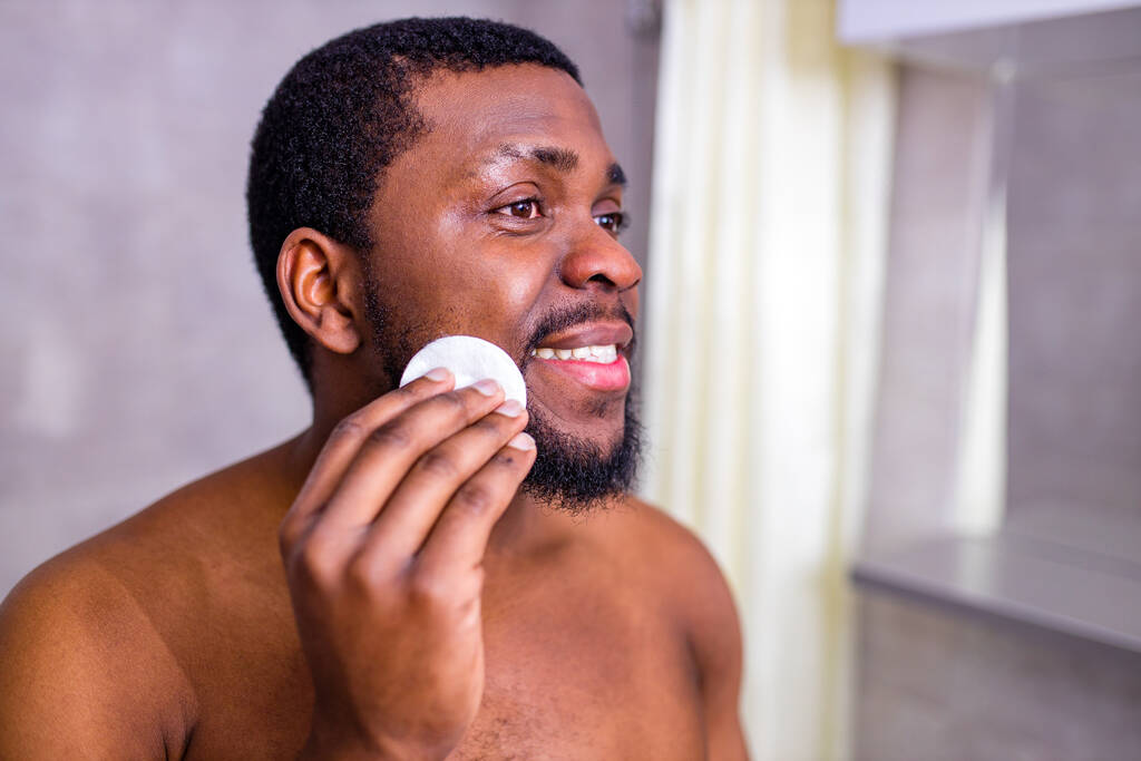 hombre afroamericano mirándose en el espejo del baño apretando espinilla - Foto, Imagen