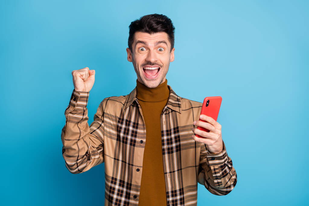 Фото молодой счастливый взволнованный сумасшедший позитивный веселый мужчина держать телефон поднять кулак в победу изолированы на синем фоне цвета - Фото, изображение