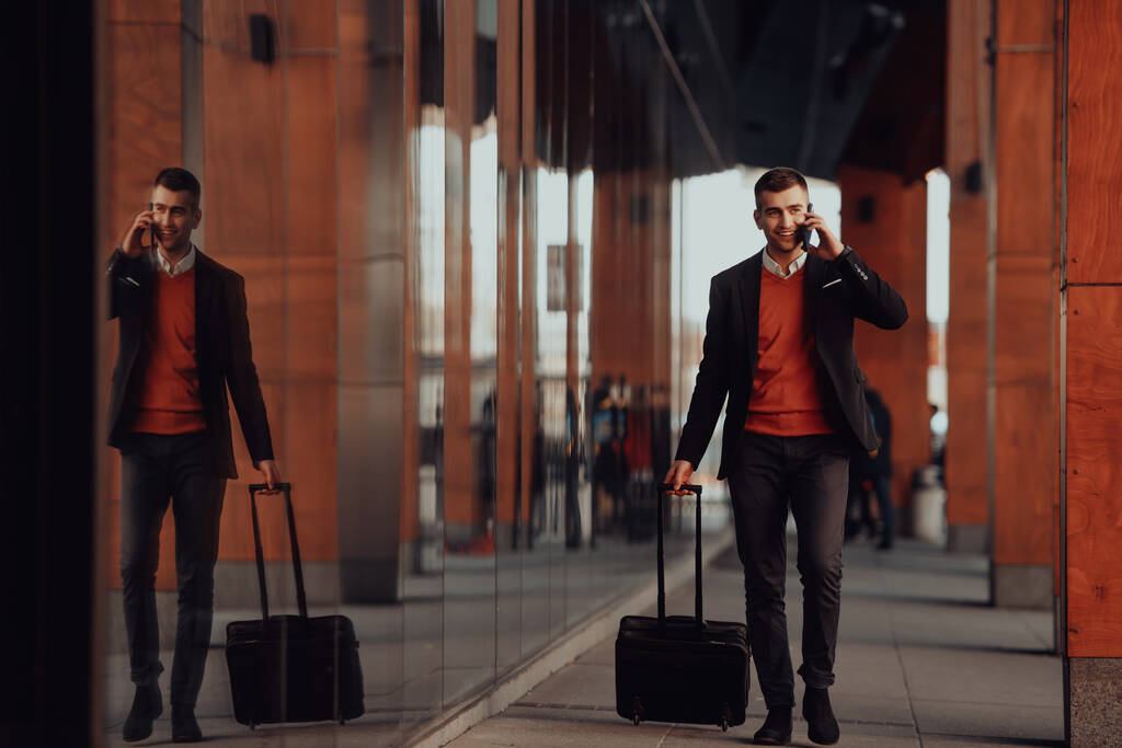 Auf zum Flughafen-Terminal. Ein junger selbstbewusster Geschäftsmann, der auf den Straßen der Stadt spaziert, seinen Koffer zieht, Kaffee trinkt und mit einem Smartphone spricht. Hochwertiges Foto - Foto, Bild