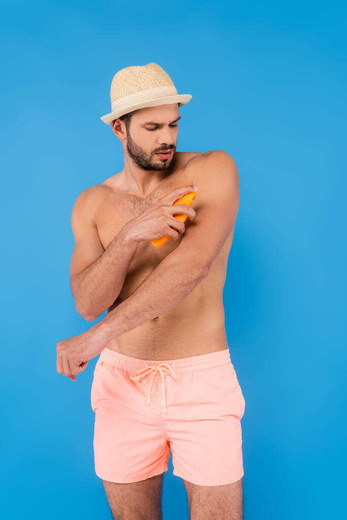 Uomo senza camicia in cappello solare applicando la protezione solare isolata sul blu  - Foto, immagini