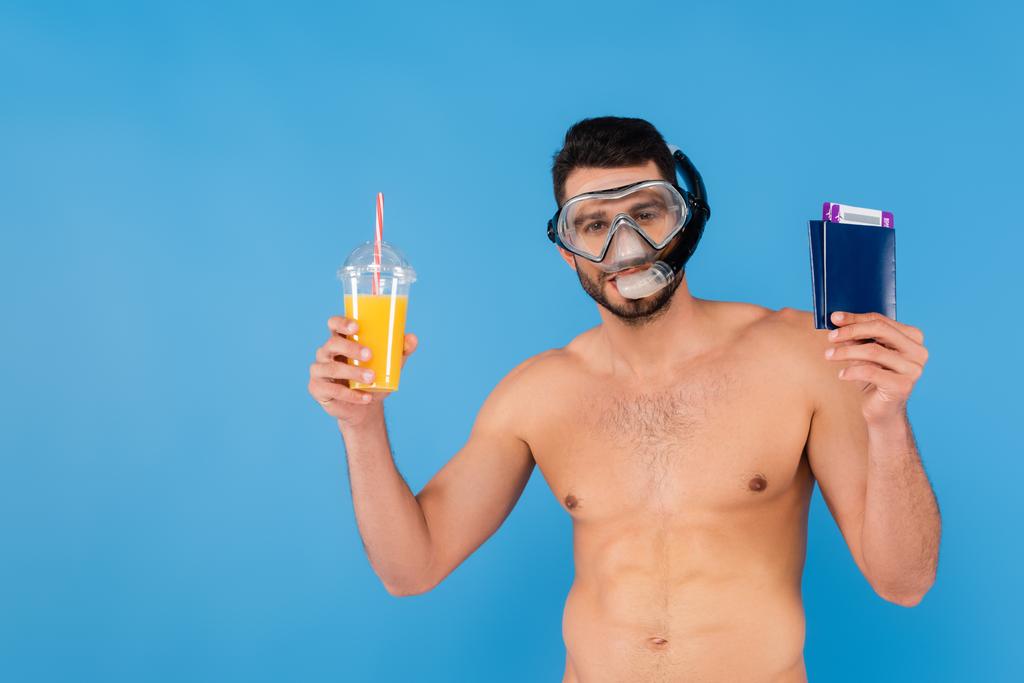 Shirtless man in swimming goggles holding passports and orange juice isolated on blue  - Photo, Image