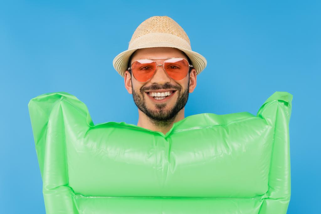 Χαμογελώντας άνθρωπος με καπέλο ήλιο και γυαλιά ηλίου κοντά σε φουσκωτό στρώμα απομονωμένο σε μπλε  - Φωτογραφία, εικόνα