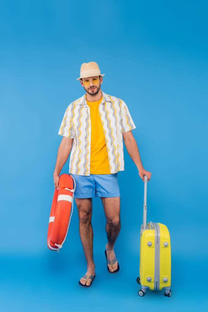 Mann mit Sonnenbrille hält Rettungsring und Koffer auf blauem Hintergrund  - Foto, Bild