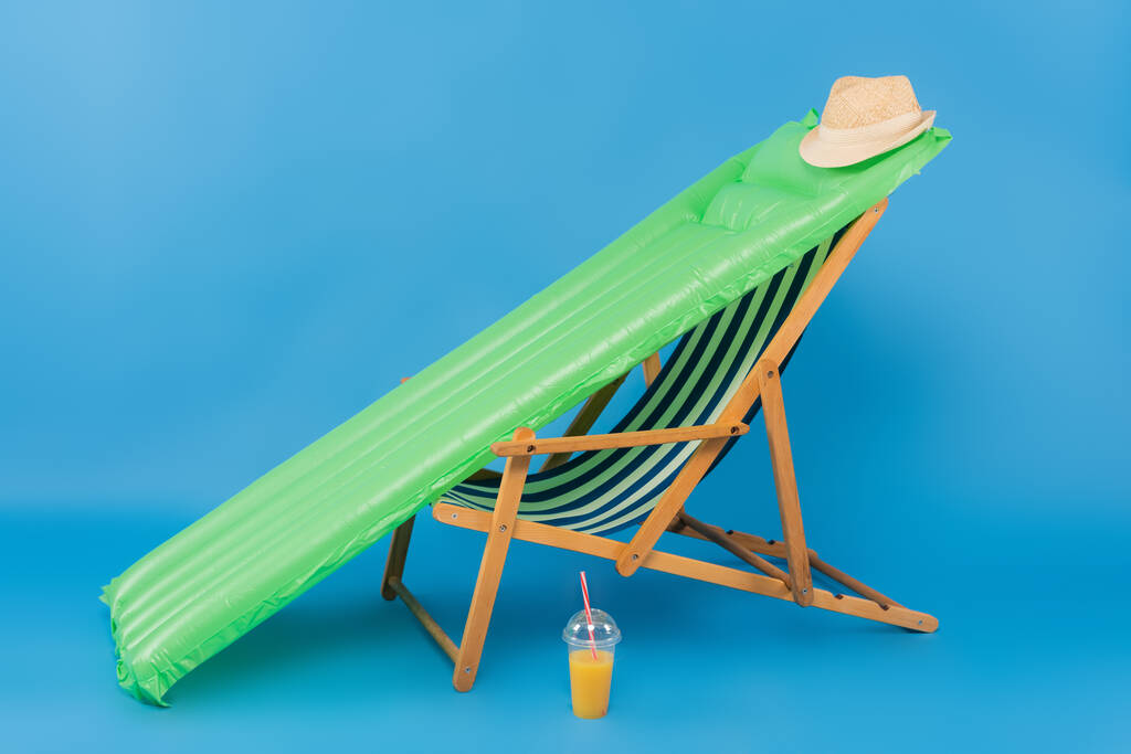 Cappello da sole su materasso gonfiabile vicino alla sedia a sdraio e succo d'arancia su sfondo blu  - Foto, immagini