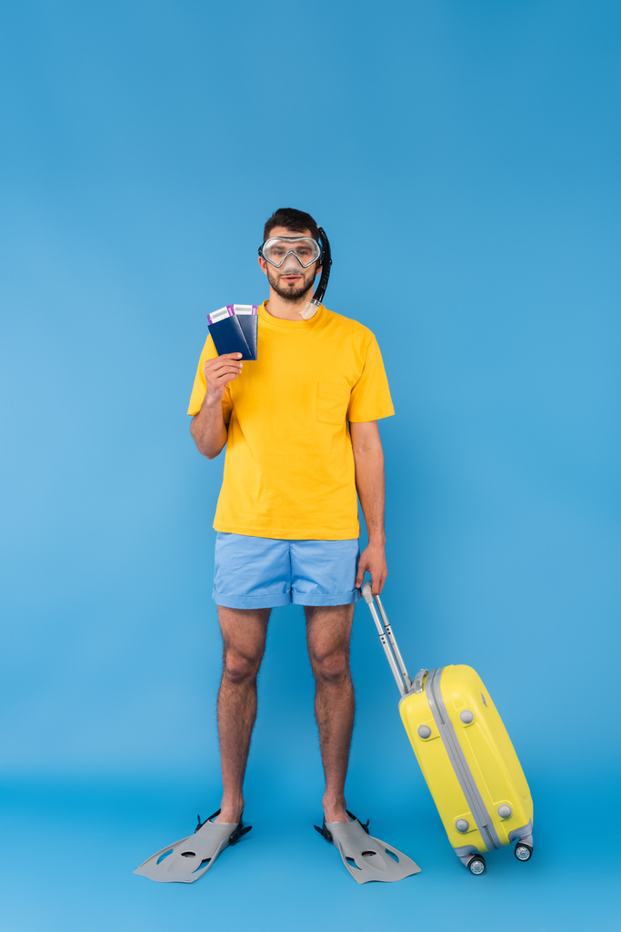 Yüzme yüzgeçli ve gözlüklü bir adam mavi arka planda pasaport ve valiz taşıyor.  - Fotoğraf, Görsel
