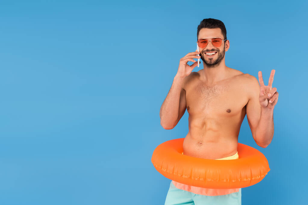 Hombre sin camisa sonriente en gafas de sol y anillo inflable hablando en el teléfono inteligente y mostrando la paz aislado en azul  - Foto, imagen