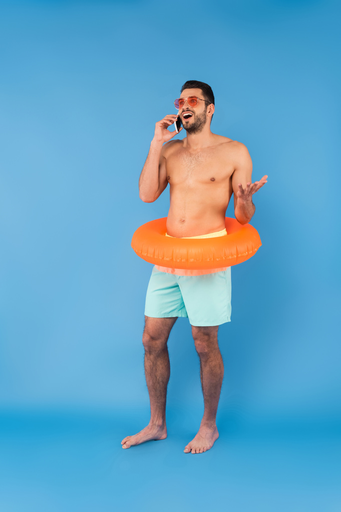 Hombre alegre sin camisa en anillo inflable y gafas de sol hablando en el teléfono inteligente sobre fondo azul  - Foto, Imagen
