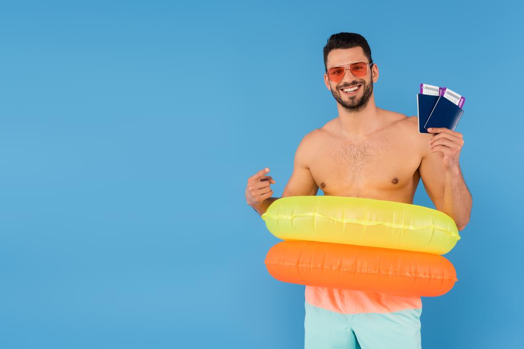 Positiver Mann mit Sonnenbrille und aufblasbaren Ringen, die auf Pässe zeigen, isoliert auf blauem Grund  - Foto, Bild