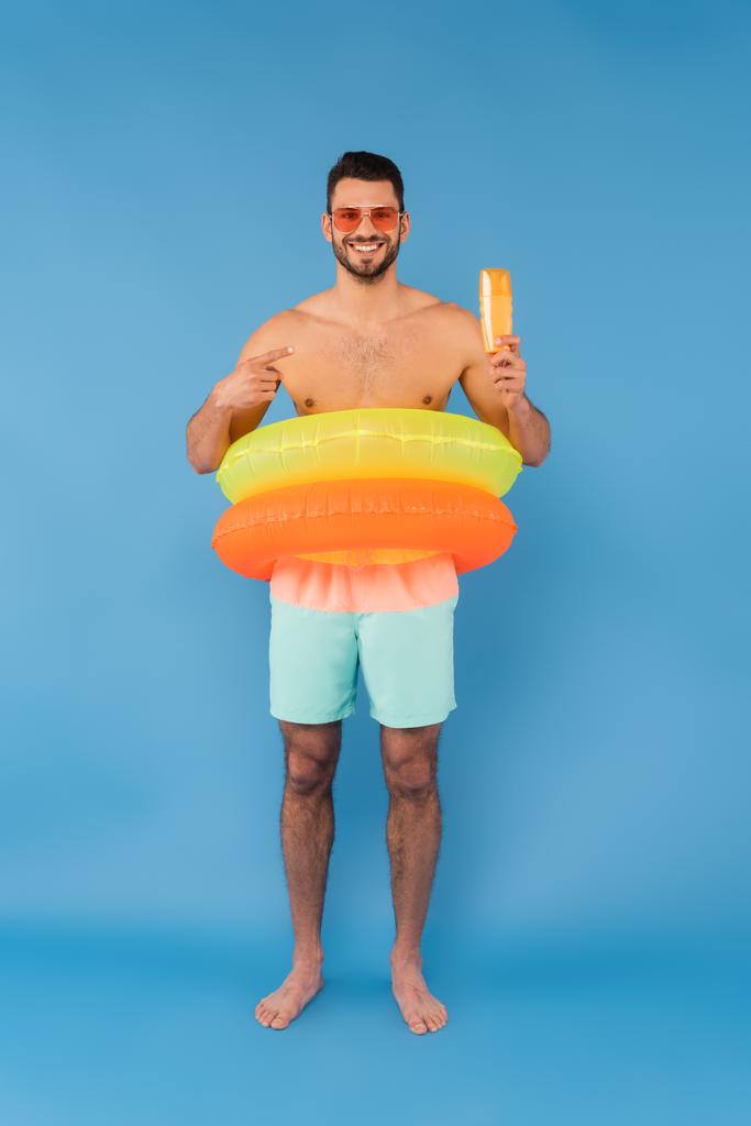 Homem sem camisa em anéis infláveis apontando para protetor solar no fundo azul  - Foto, Imagem
