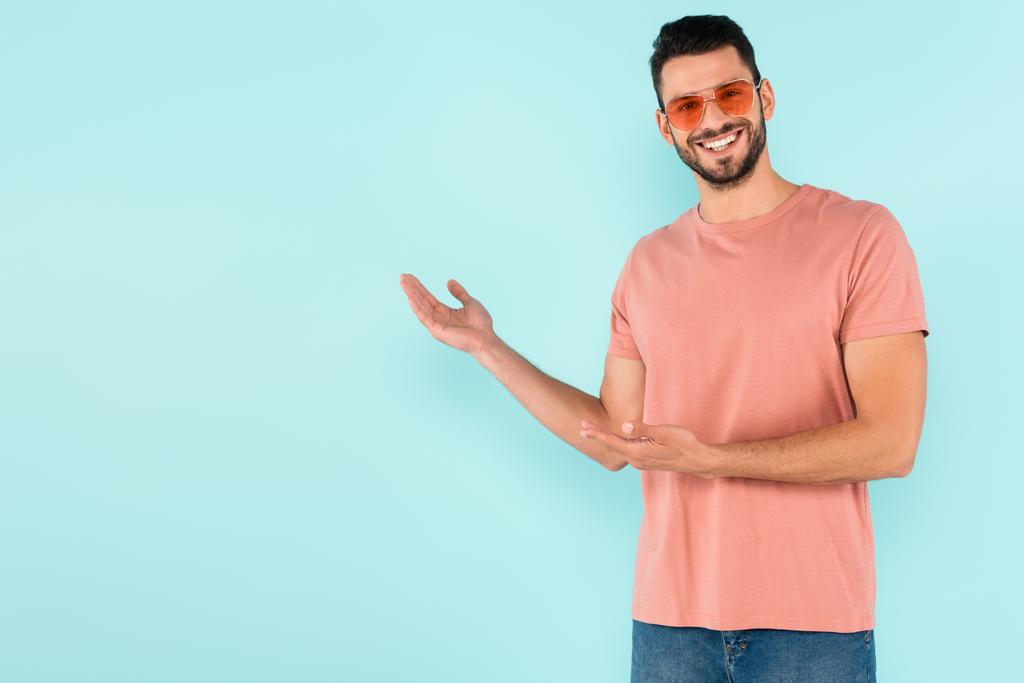 Lächelnder Mann mit Sonnenbrille zeigt mit den Händen auf blauem Grund  - Foto, Bild
