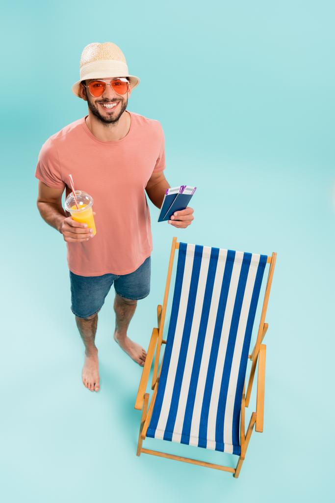 Wysoki kąt widzenia pozytywnego człowieka w okularach przeciwsłonecznych trzyma sok pomarańczowy i paszporty w pobliżu leżaka na niebieskim tle  - Zdjęcie, obraz