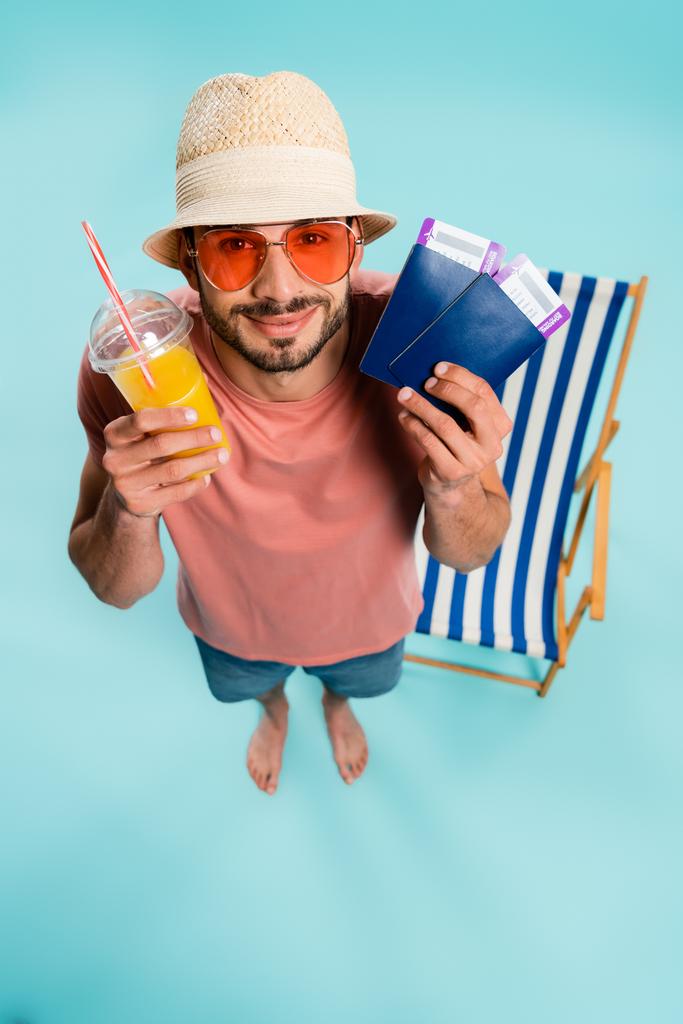 Vue du dessus de l'homme en lunettes de soleil tenant le jus d'orange et passeports avec des billets d'avion près de chaise longue floue sur fond bleu  - Photo, image