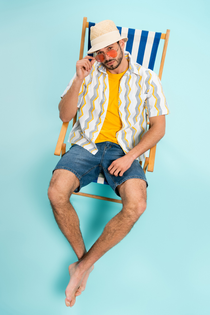 Vue grand angle de l'homme barbu en lunettes de soleil et chapeau de paille regardant la caméra sur fond bleu  - Photo, image