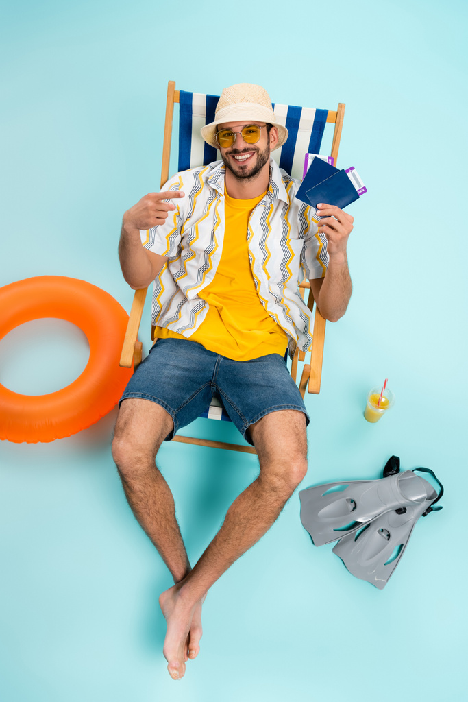 Vista de ángulo alto del hombre sonriente apuntando a pasaportes cerca de aletas de natación, jugo de naranja y anillo inflable sobre fondo azul  - Foto, imagen