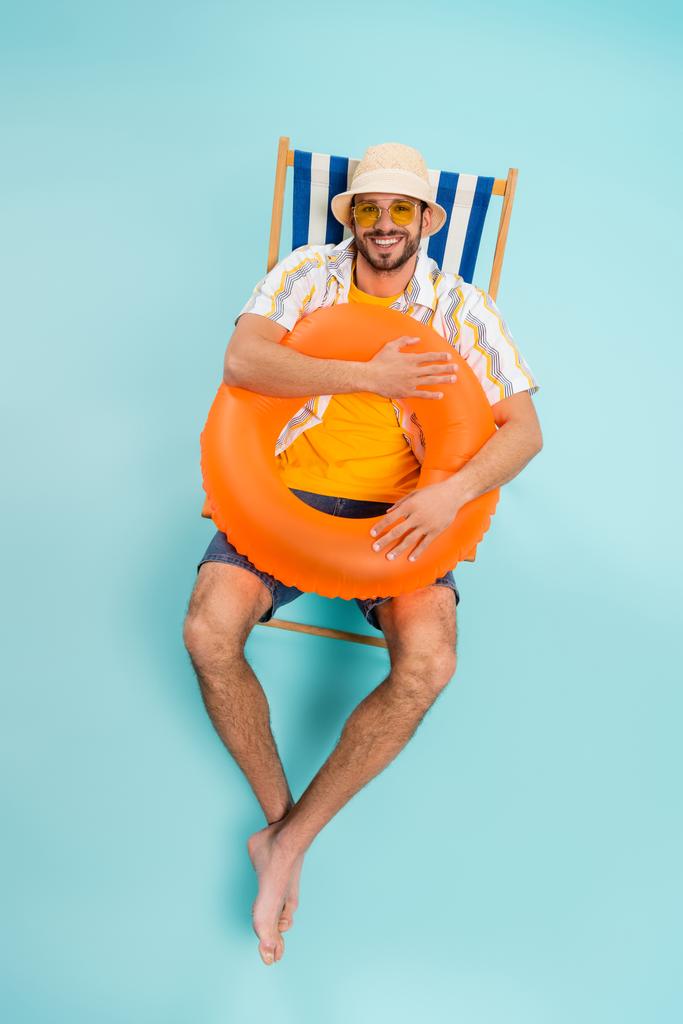 Υψηλή γωνία άποψη του θετικού ανθρώπου σε ψάθινο καπέλο κρατώντας φουσκωτό δαχτυλίδι στο κατάστρωμα καρέκλα σε μπλε φόντο  - Φωτογραφία, εικόνα