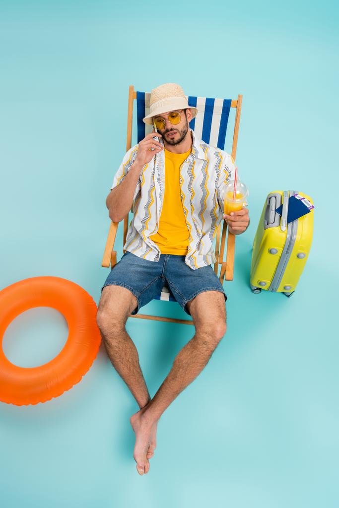 男性の高い角度ビューでオレンジジュースとスマートフォンで話す近くインフレータブルリングとスーツケース上の青の背景  - 写真・画像