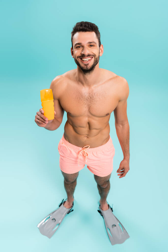 Hoge hoek uitzicht van shirtloze man in zwemvliezen met zonnebrandcrème op blauwe achtergrond  - Foto, afbeelding