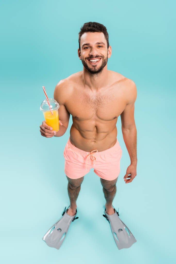 Visão de alto ângulo do homem sem camisa feliz em nadadores segurando suco de laranja no fundo azul  - Foto, Imagem