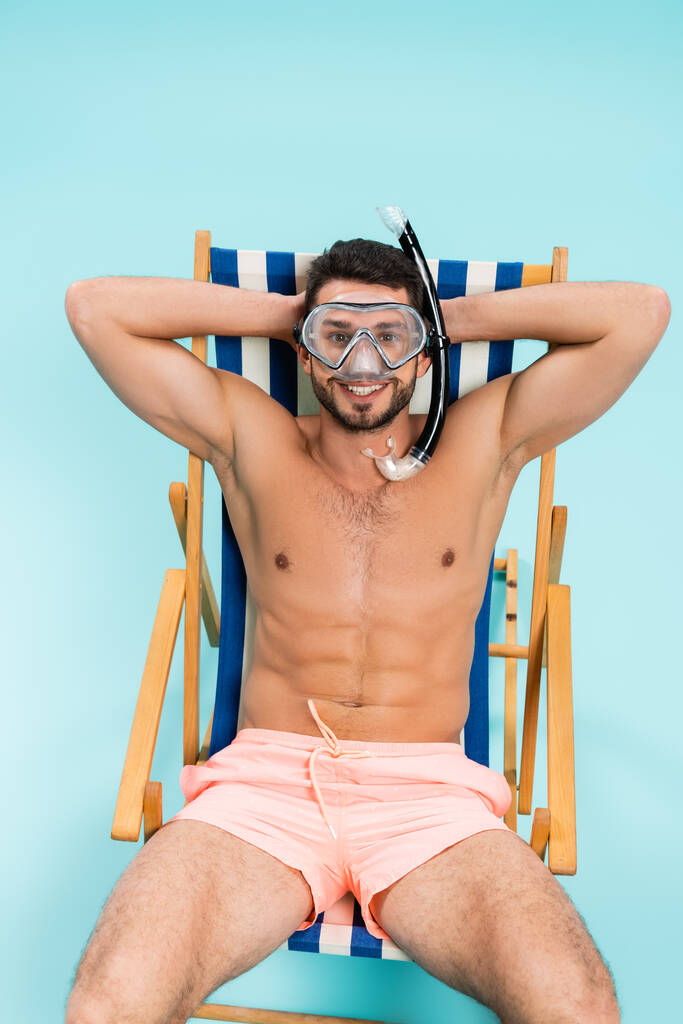 Pozitivní muž bez trička v plaveckých brýlích sedí na palubě židle na modrém pozadí  - Fotografie, Obrázek