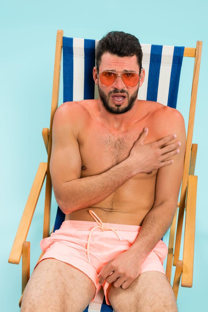 Opgewonden man in zonnebril raakt zonnebrand op dek stoel geïsoleerd op blauw  - Foto, afbeelding