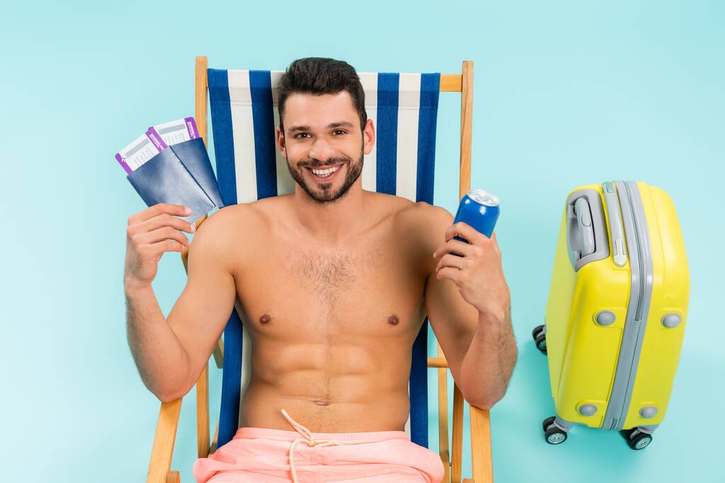 Usmívající se muž bez košile držící konzervované nápoje a pasy v blízkosti kufru na modrém pozadí  - Fotografie, Obrázek