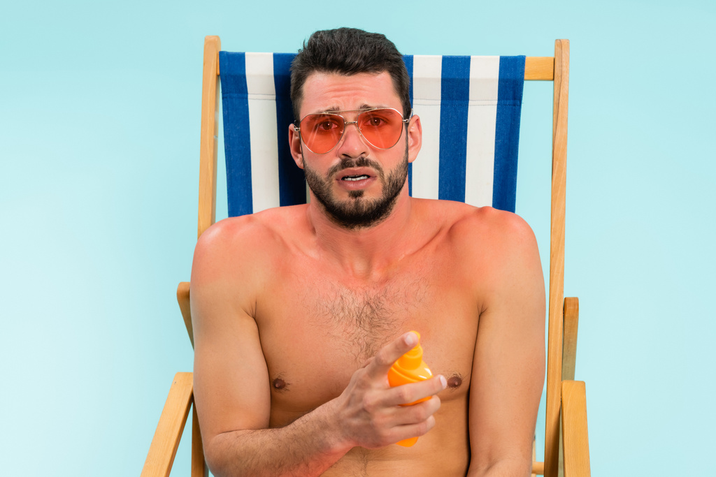 Besorgter Mann mit Sonnenbrand hält Sonnencreme auf Liegestuhl isoliert auf blauem Grund  - Foto, Bild