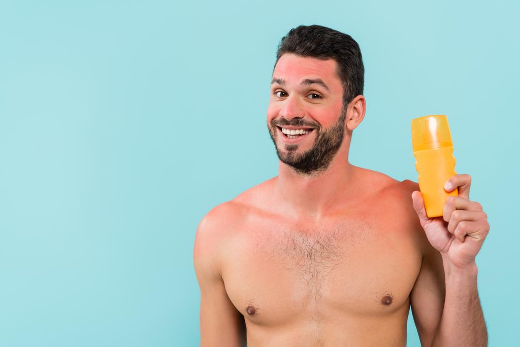 Shirtless man with sunburn holding sunscreen and smiling isolated on blue  - Photo, Image