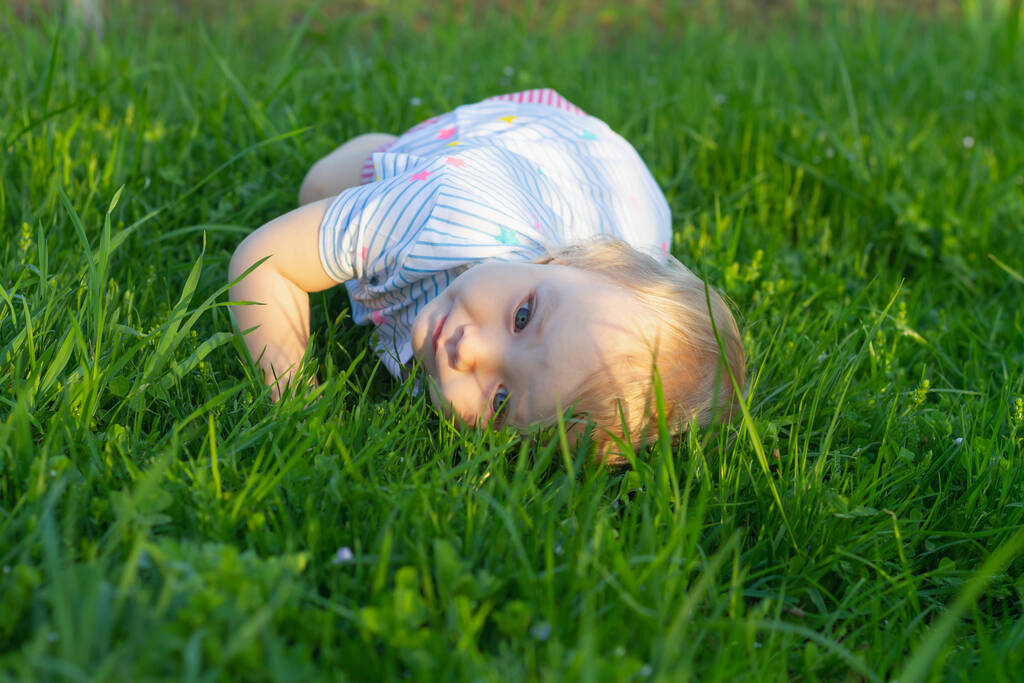 Όμορφο κοριτσάκι 2 ετών ξαπλώνει στο γρασίδι το καλοκαίρι και χαμογελά. - Φωτογραφία, εικόνα