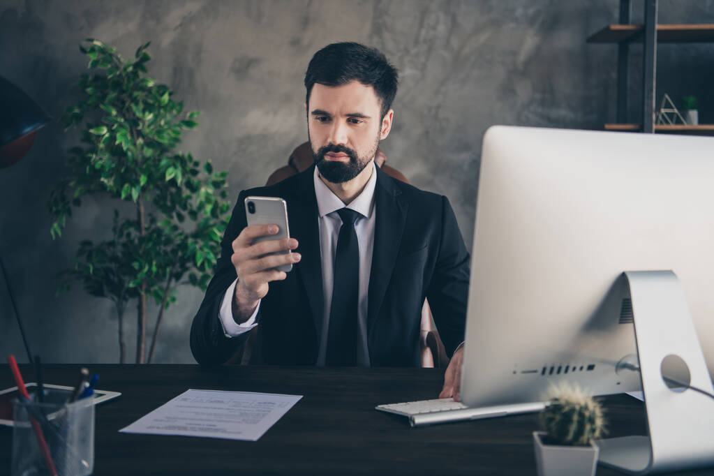 Foto de hombre exitoso sentarse escritorio ordenador sostener teléfono mirar pantalla leer sms usar traje camisa corbata en la oficina moderna en el interior - Foto, imagen
