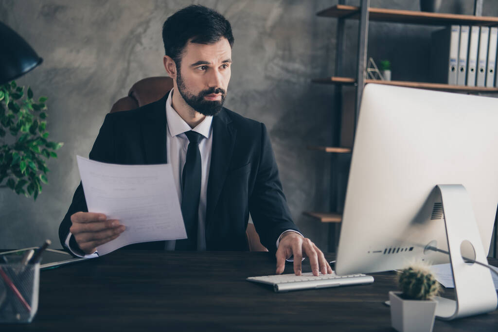 Фото сосредоточенного босса сидеть настольный компьютер смотреть экран держать отчет бумажный костюм галстук галстук в современном офисе в помещении - Фото, изображение