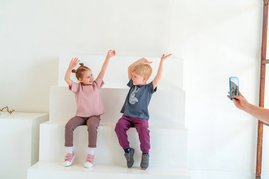 Kleine gelukkige jongen en meisje in modieuze kleding glimlach, poseren tegen witte muur studio, zittend op stappen. concept reclame voor kinderkleding, emotionaliteit kinderen, het dynamisme beeld. Kopieerruimte - Foto, afbeelding