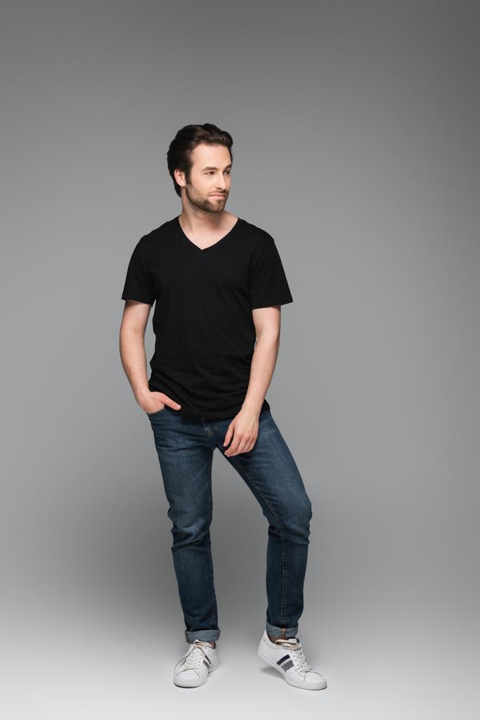 volledige lengte van de bebaarde man in jeans en zwart t-shirt poseren met de hand in zak op grijs - Foto, afbeelding