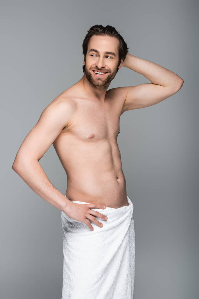glücklicher und muskulöser Mann in weißes Handtuch gehüllt, stehend mit der Hand an der Hüfte isoliert auf grau - Foto, Bild