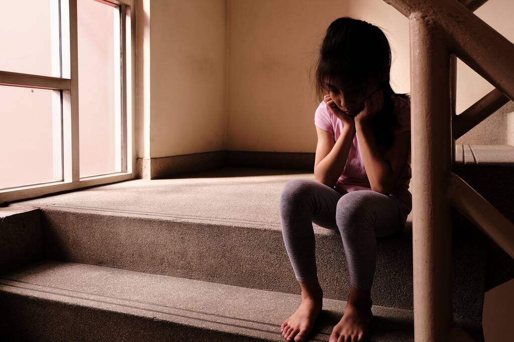 Koncepcja wykorzystywania dzieci. Smutna i samotna młoda dziewczyna siedząca przy schodach wewnątrz budynku ze światłem przez szklane okno w tle. - Zdjęcie, obraz