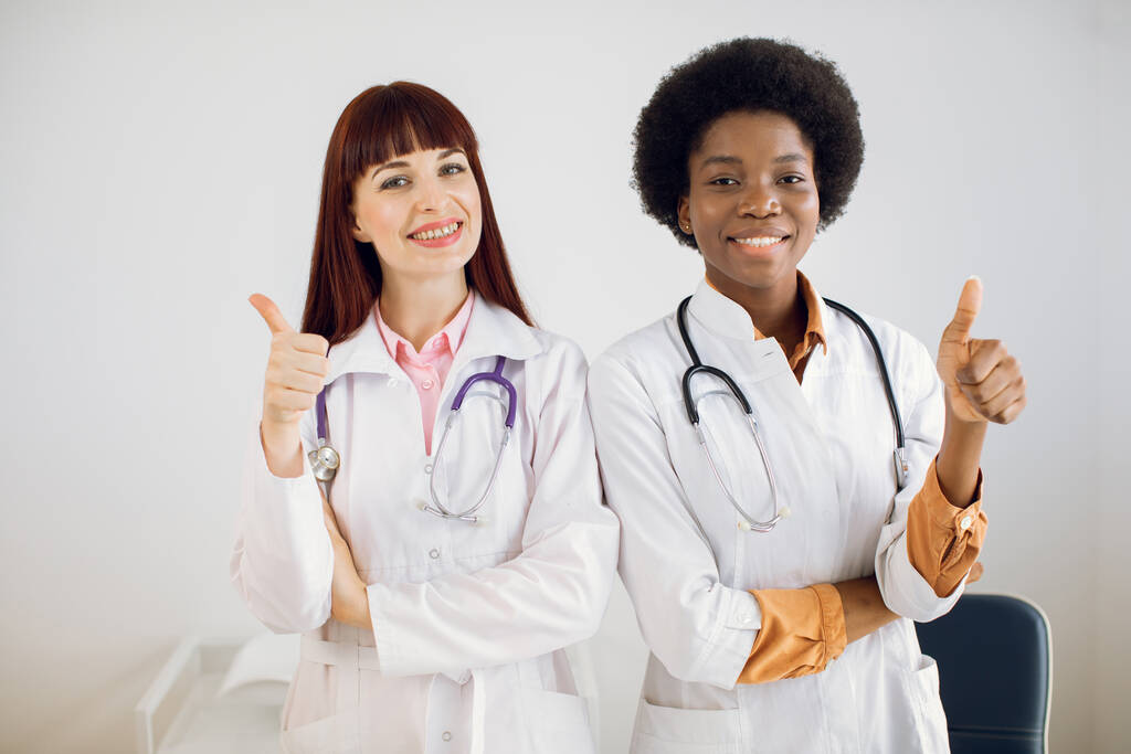 医療援助、保険、医療、医療の概念。病院のオフィスで親指を立てたチームの一員として二人の多人種の女性医師がカメラに向かって笑っています. - 写真・画像