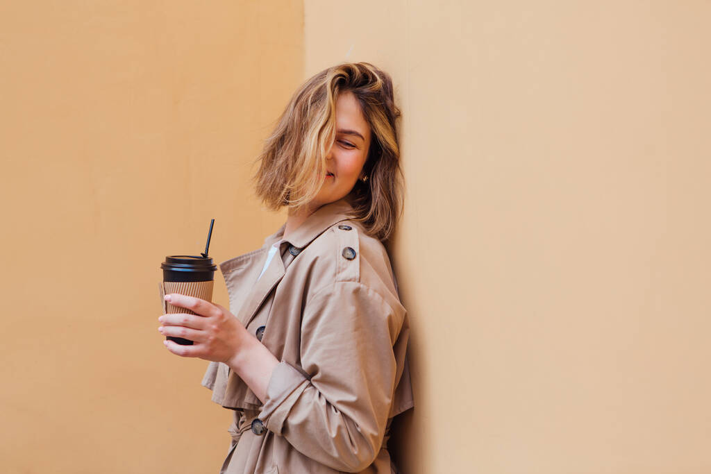 Fiatal, mosolygós, évezredes nő, vad hajjal, őszi kabátban, egy csésze kávé mellett, hogy a bézs fal közelébe mehessen.. - Fotó, kép