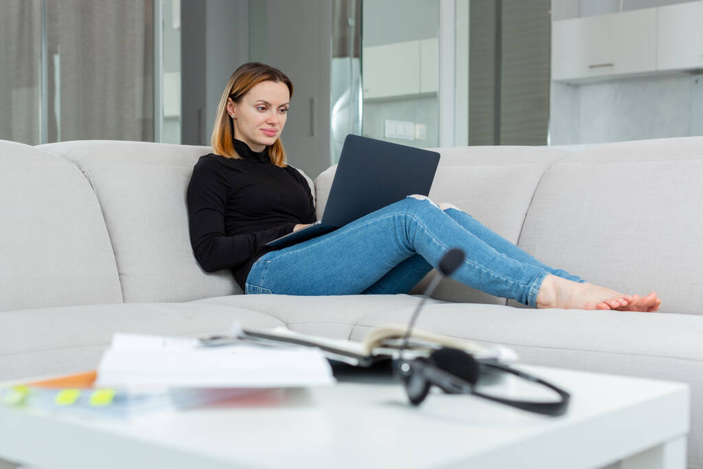 Attraktives Mädchen arbeitet von zu Hause aus hinter einem Laptop und sitzt auf der Couch - Foto, Bild