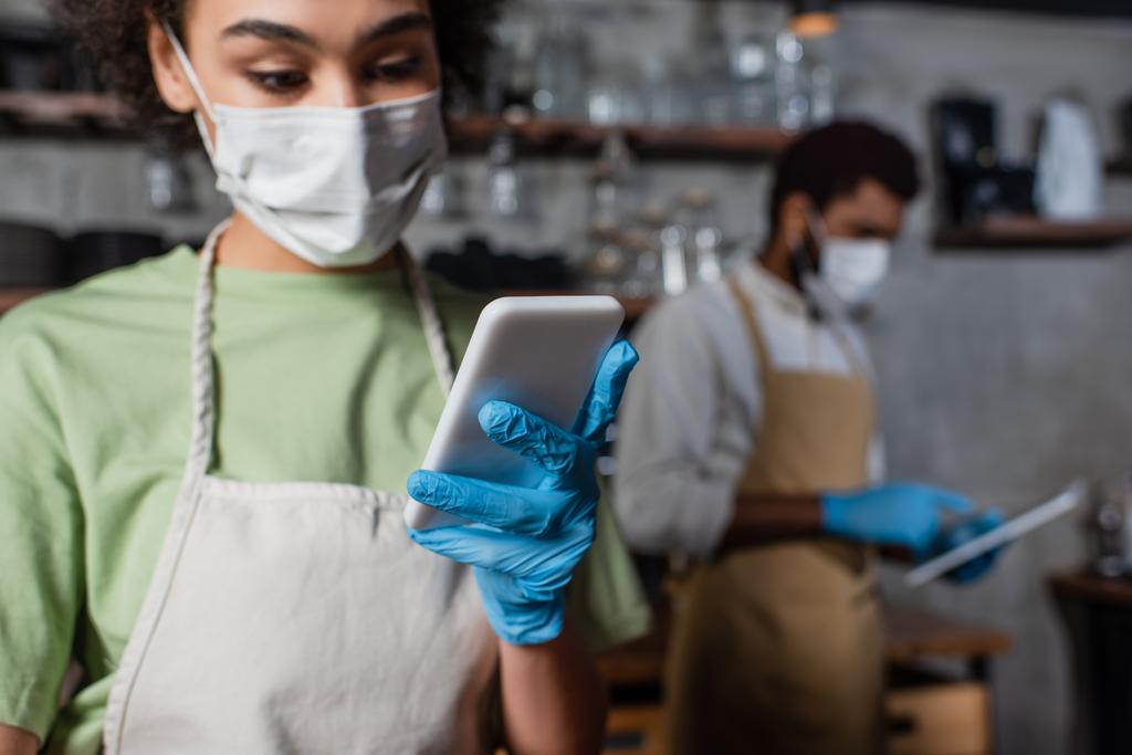 Smartphone in mano di barista afroamericano in maschera medica su sfondo sfocato  - Foto, immagini