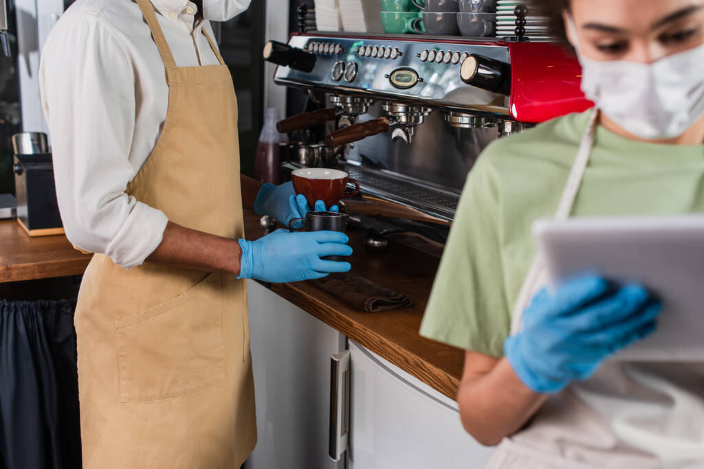 Barista afro-américaine en gants de latex tenant la tasse près de la machine à café et collègue flou en utilisant une tablette numérique  - Photo, image