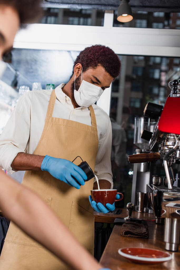 Afrikai amerikai barista orvosi maszkban és latex kesztyűben kávét készít elmosódott kollégája közelében a kávézóban  - Fotó, kép