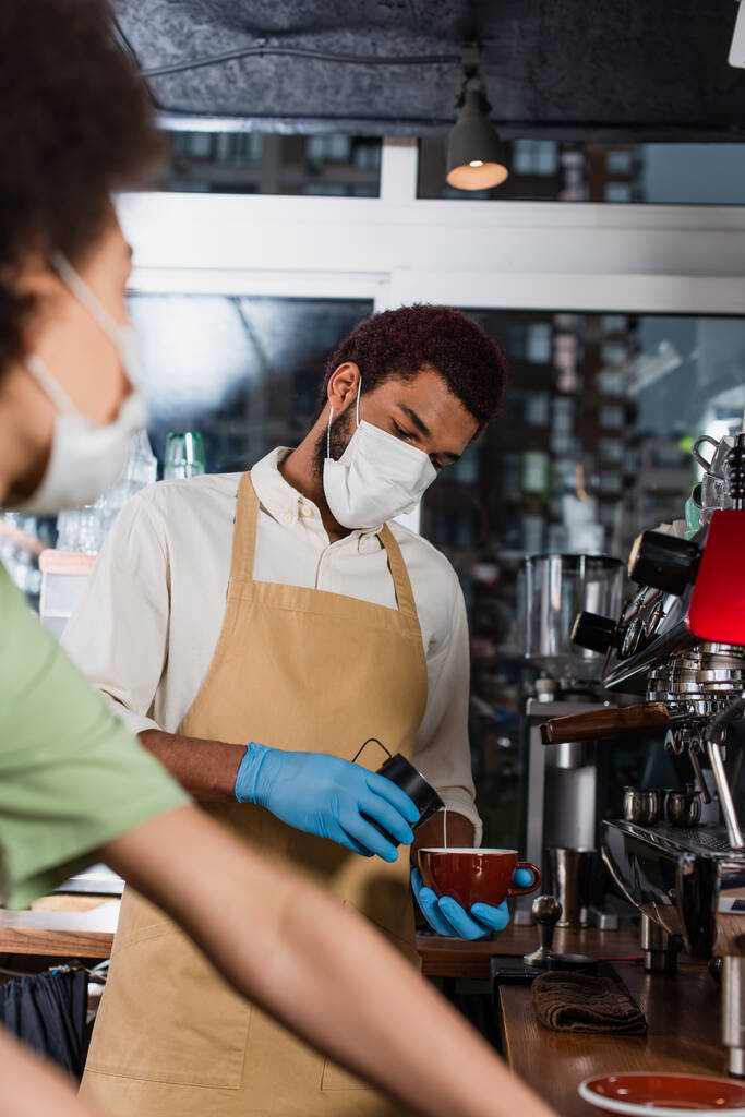 Afro-Amerikaanse barista in beschermende masker en latex handschoenen het maken van koffie in de buurt wazig collega  - Foto, afbeelding