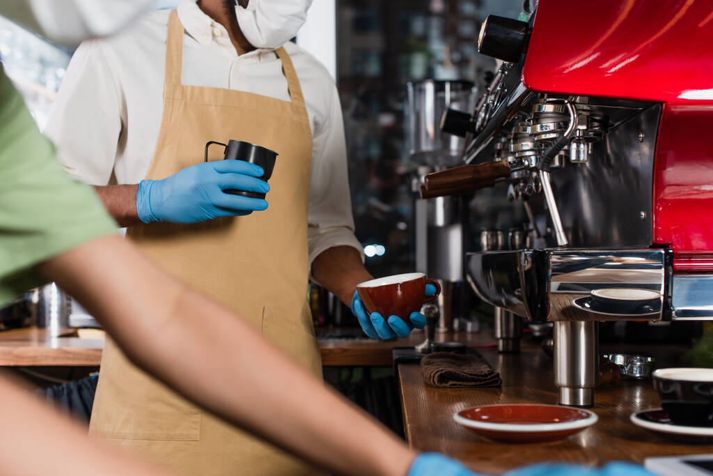 Gewassen beeld van Afrikaanse Amerikaanse barista in medisch masker en latex handschoenen met kopje en het maken van koffie in de buurt wazig collega  - Foto, afbeelding