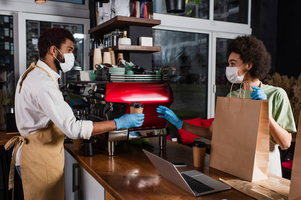 Afrikai amerikai baristák orvosi maszkban és latex kesztyű kezében kávé menni, és papírzacskó kávézóban  - Fotó, kép