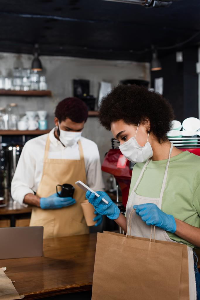 Afro-Amerikaanse barista in medisch masker met behulp van smartphone en het houden van papieren zak in de buurt wazig collega  - Foto, afbeelding