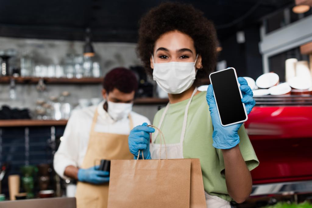 Smartphone en mano de barista afroamericano en máscara médica sosteniendo bolsa de papel sobre fondo borroso  - Foto, imagen