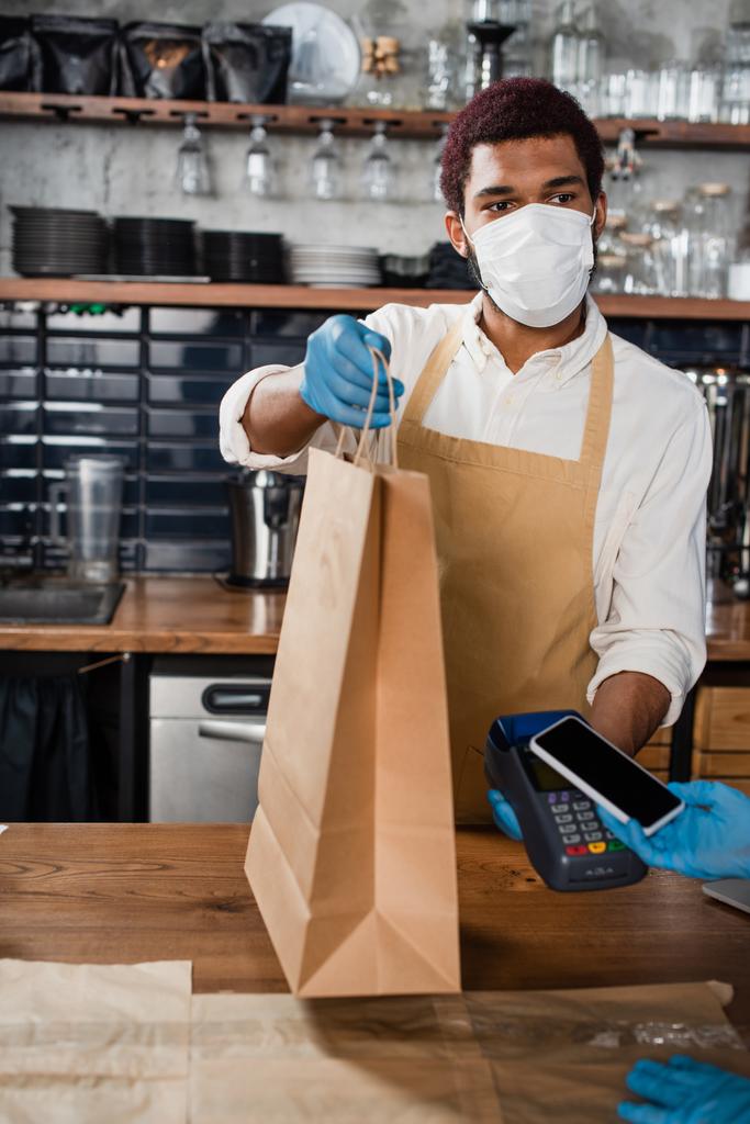 Afrykański amerykański barista w masce medycznej trzymając papierową torbę i terminal płatniczy w pobliżu klienta ze smartfonem  - Zdjęcie, obraz